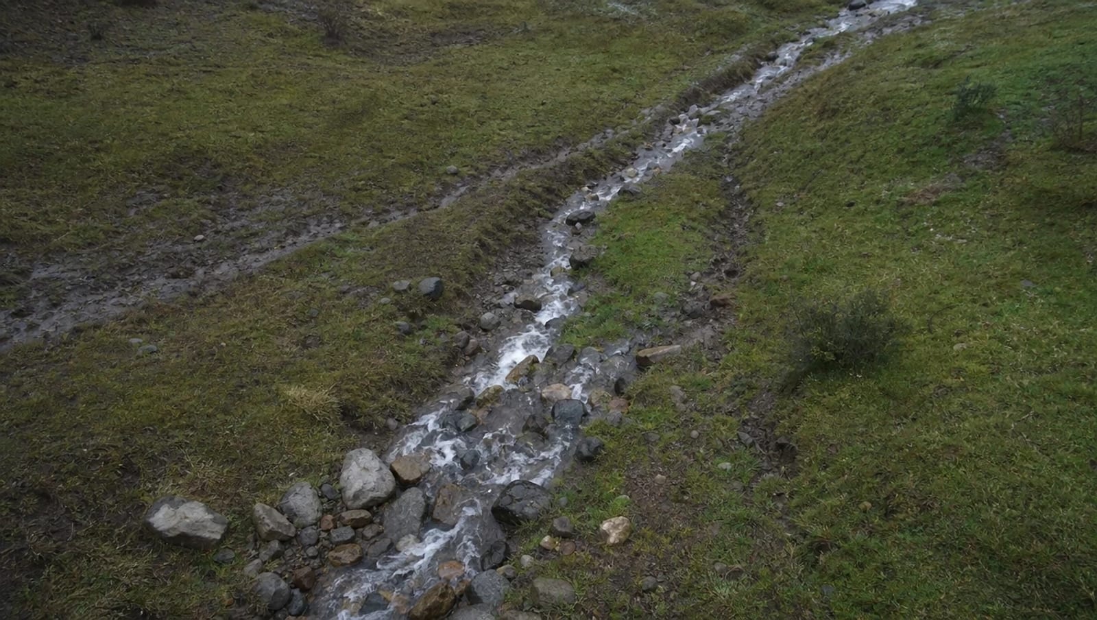Escurrimiento de agua entre piedras atravesando un terreno con pasto, con la huella del cauce marcada en el suelo